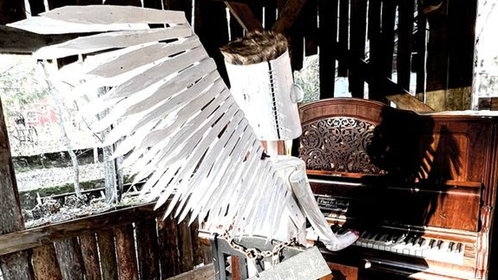 Kuvassa on tummassa raakapuusta rakennetussa tilassa vanhanaikaista pianoa soittamassa valkoisista siivistä tehty hahmo, jolla on torven muotoiset valkoiset käsivarret ja kädet soittamassa sekä valkoinen tötteröpää.