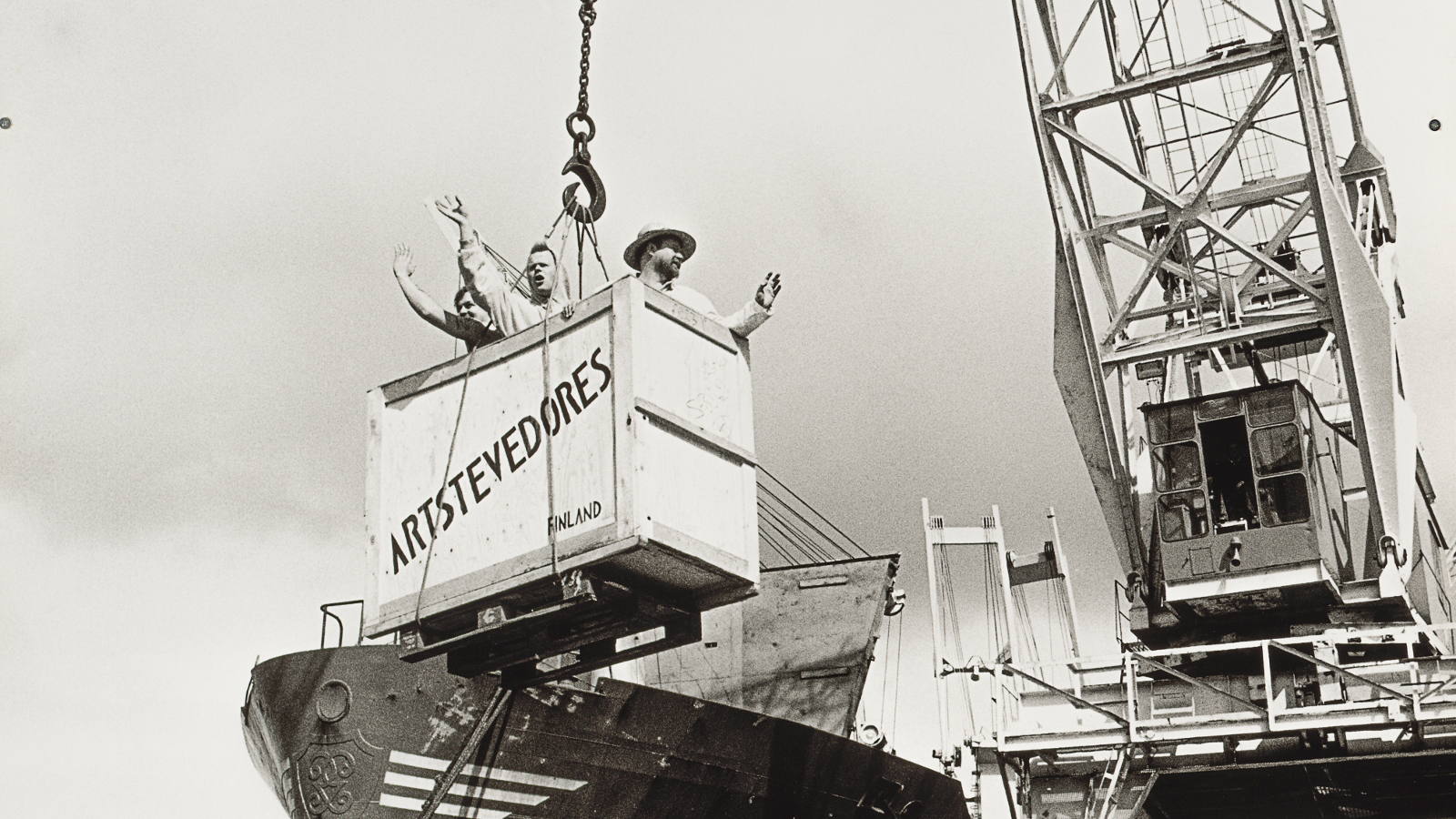 Kuvassa on satamanosturi laskemassa puulaatikkoa laivan kannelle. Laatikossa on teksti Artstevendores. Kuva on musta-valkoinen.