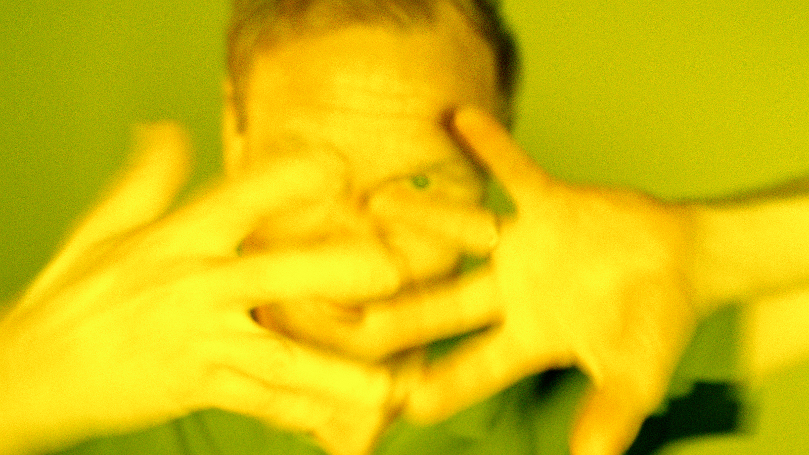 Kuvassa on Ismo Alangon kasvot ja ne ovat edessä olevien käsien takana. Sormet on haristettu. Kuvan sävy on vihertävä.