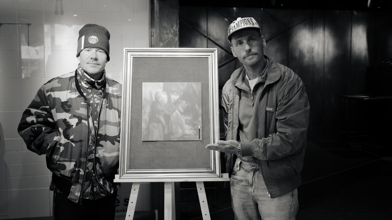 Kuvassa ovat vasemmalla VilleGalle ja oikealla Jare. Heidän välissään on raamitettu taulu varustettuna kolmella jalalla. Kuva on musta-valkoinen.