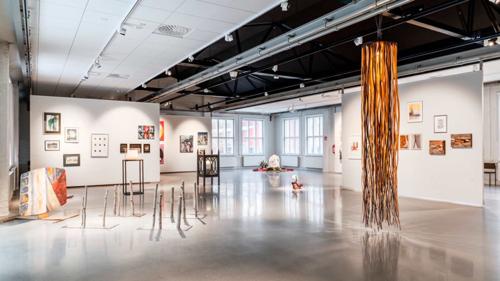Kuvassa on vaalean harmaat seinät taidenäyttelytilassa ja siinä on eri kokoisia taideteoksia seinillä ja takana yksi pylväs.