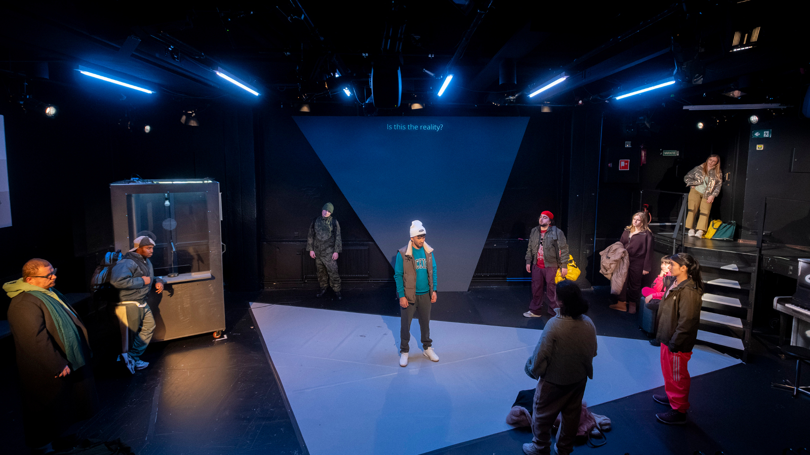 Kuvassa on näyttämö, johon kohdistuu sinistä valoa. Keskellä seisoo nuori mies talvivaatteissa. Ympärillä kauempana on joka puolella näyttämön reunalla nuoria ulkovaatteissa.