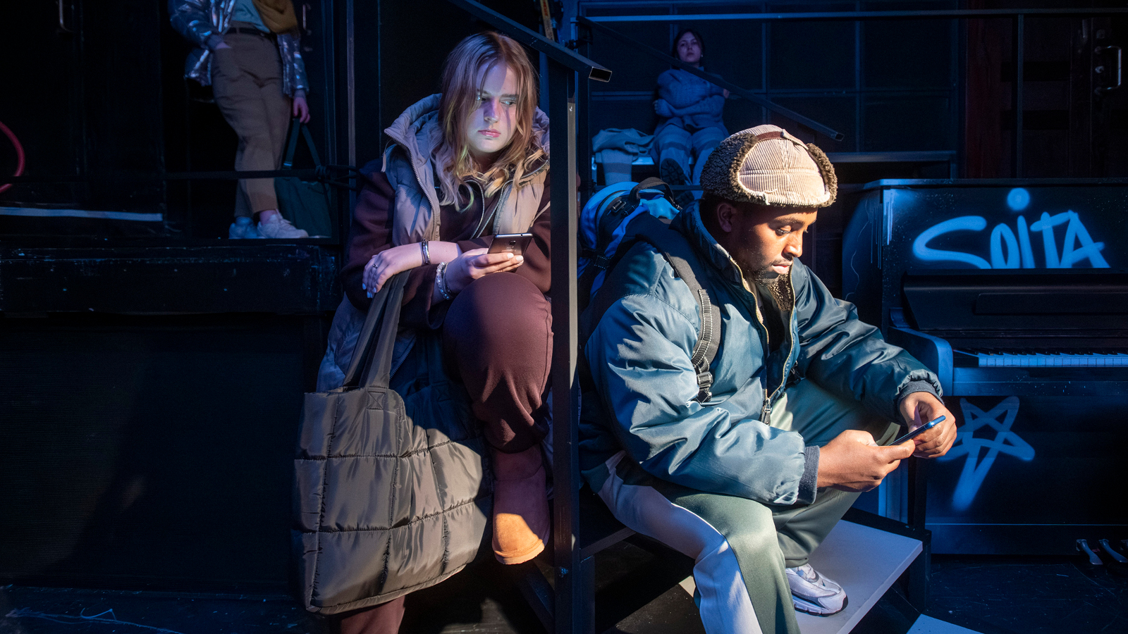 Kuvassa on oikealla nuori mies istumassa karvahatussa ja toppatakissa. Hänen takanaa vasemmalla on nuori nainen, jonka hiukset roikkuva ja hän nojaa vasempaan jalkaansa. Tausta on osittain tumma.