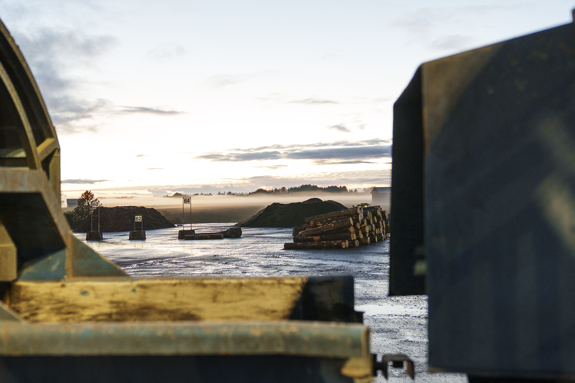 Kuvassa on vasemmalla kellertäviä mrtallirakenteita. Edessä on vettä ja kauempanapuupino. Oikealla on tumma, iso metallinen seinämä.