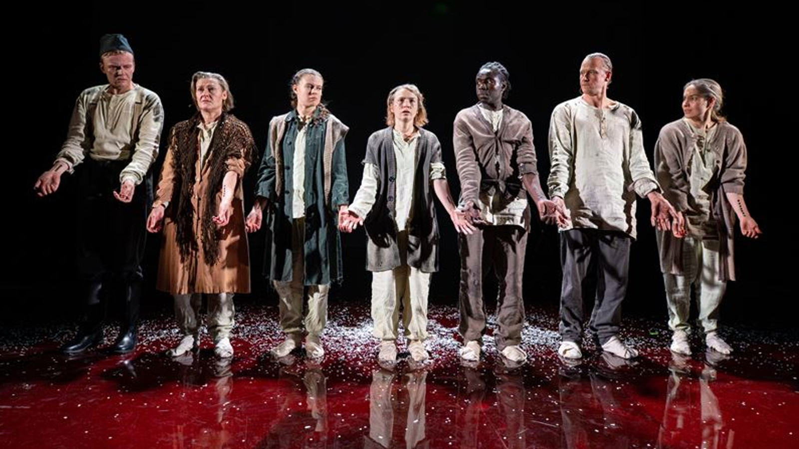 Kuvassa on seitsemän henkilöä rivissä kauhtuneissa takeissa ja vaaleissa puseroissa. Tausta on tumma ja jalkojen alla on vaaleita hiutaleita.