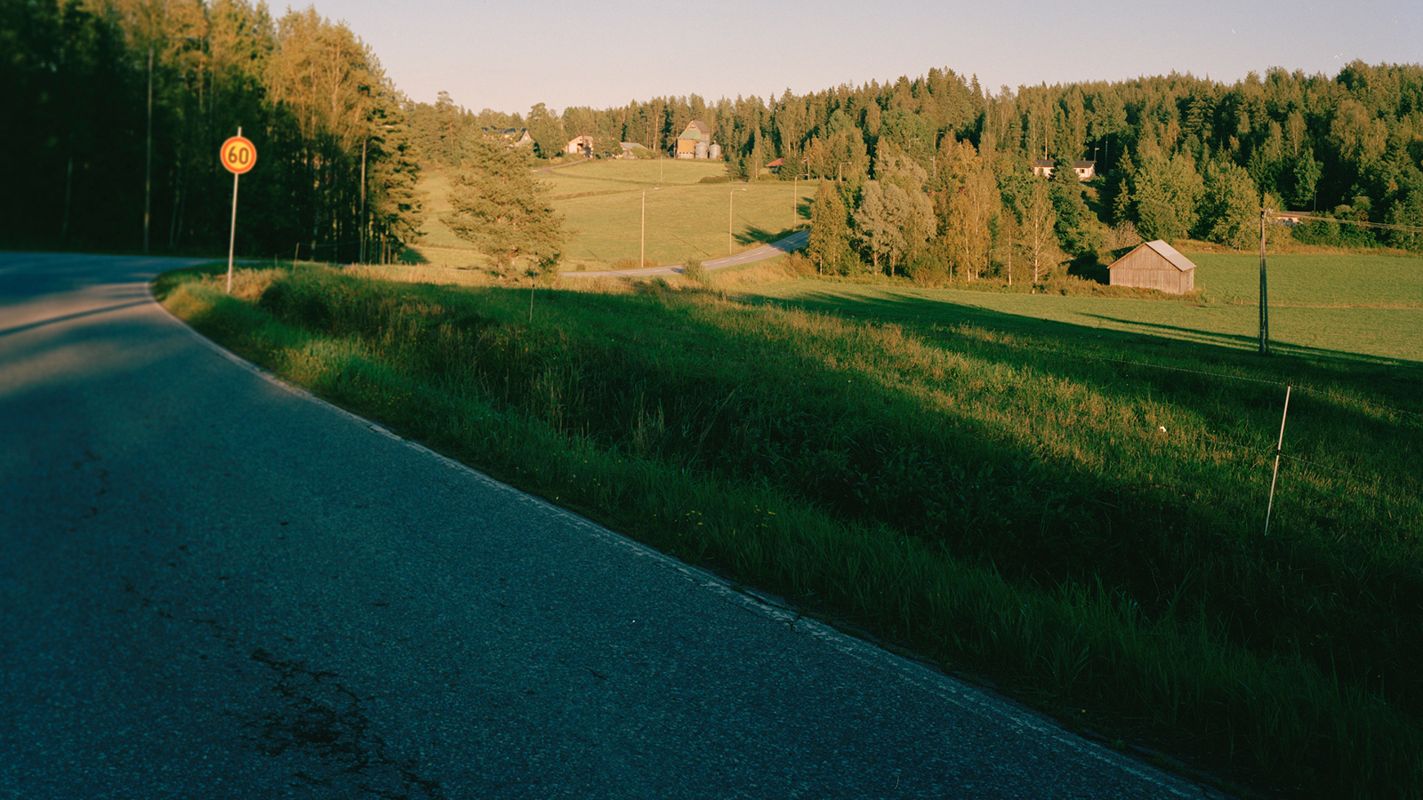 Kuvassa on vasemmalla varjossa kaartuva tie maalaismaisemassa. Taustalla näkyy pienen laakson takana metsää, peltoa ja taloja.