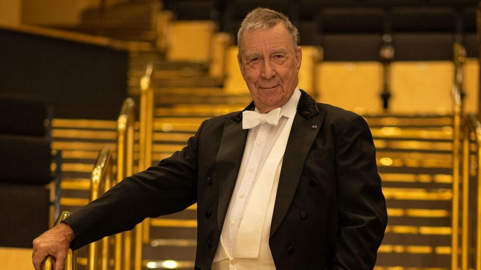 Kuvassa on Okko Kamu seisomassa portaikossa, jossa on metalliset kaiteet. Hänellä on frakki päällään ja pitää oikeata kättään kaiteella. Kuvan sävy on ruskehtava.