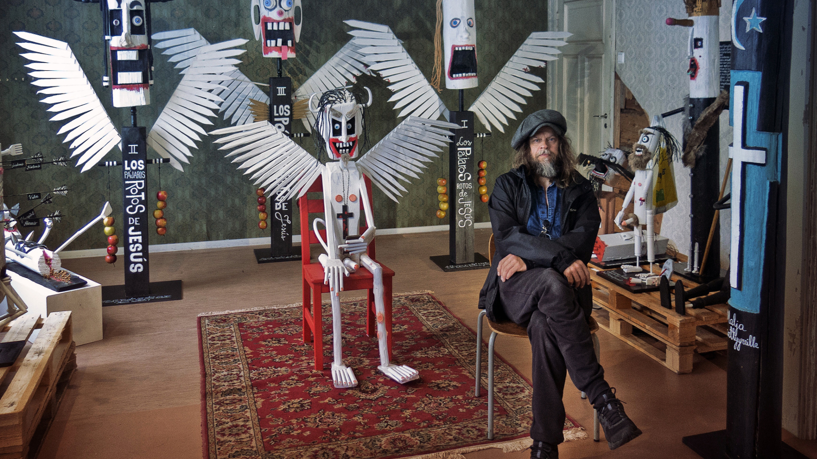 Kuvassa on Jori Tapio Kalliola istumassa tuolilla etuoikealla.  Taustalla on hänen näyttelytilaansa.  Siellä on värikkäitä töitä ja metallisia linnunsiipisiä olentoja keskellä.