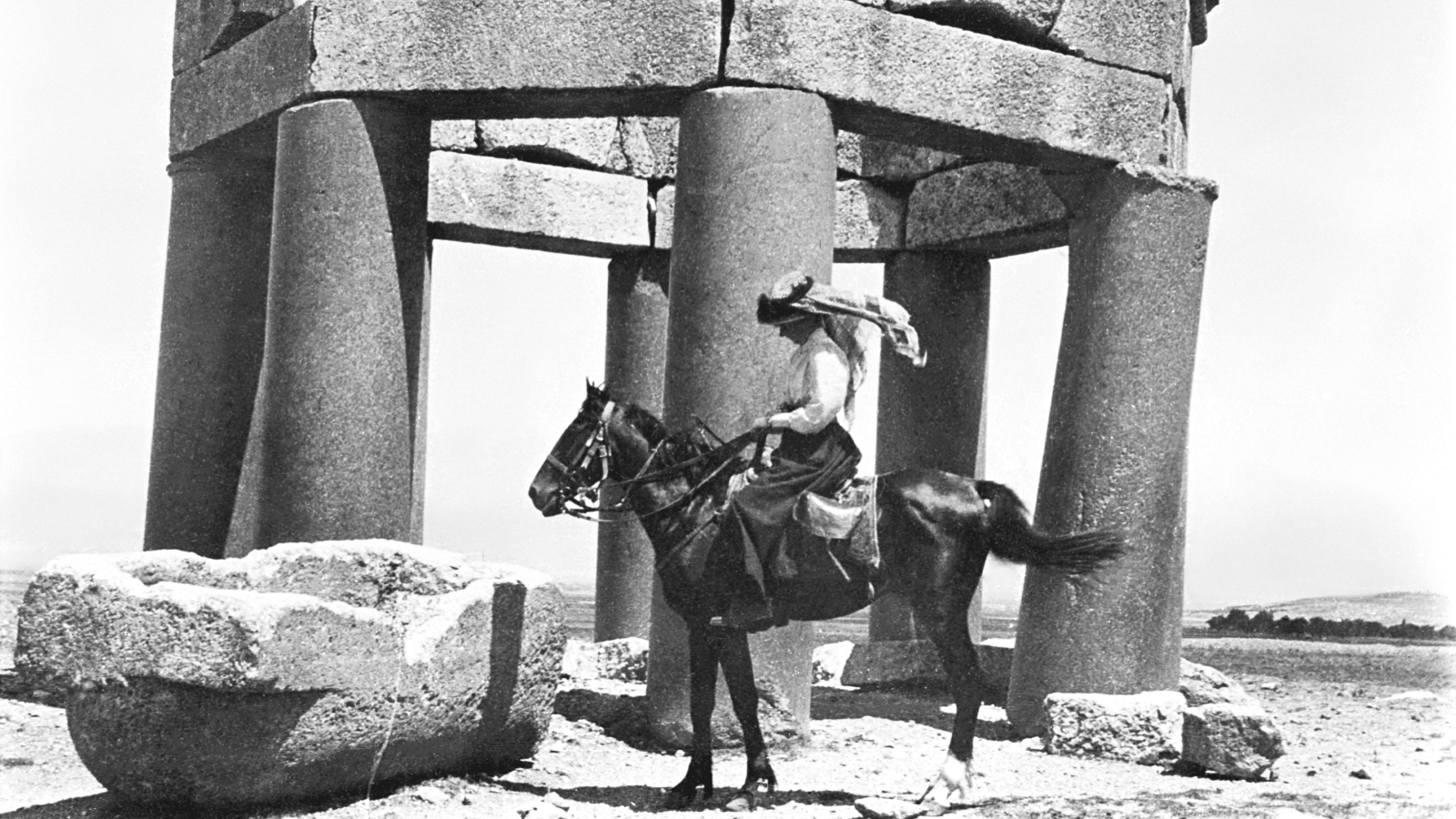 Kuvassa on ratsastaja, jolla on arabipäähine.  Taustalla on isoista kivipylväistä tehty kuusikulmainen rakennelma ja edessä on kivinen vesiallas.  Kuva on musta-valkoinen.