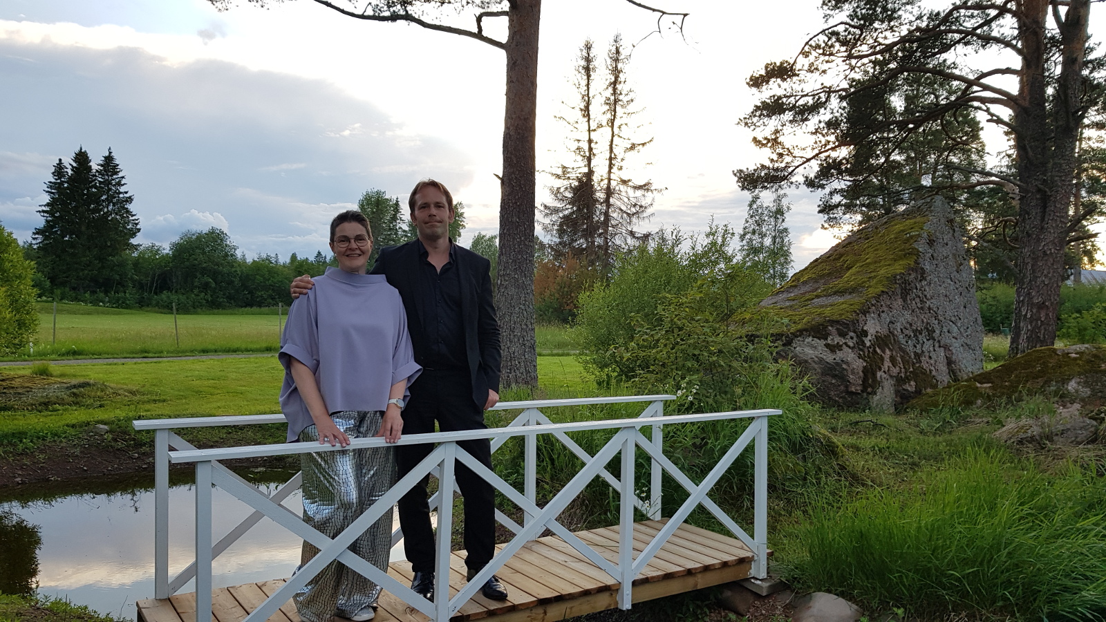 Kuvassa ovat Siru Ahopelto ja Jukka Merjanen seisomassa valkoisella sillalla, joka menee puron yläpuolella. Siru Ahopellolla on vaalean harmaa pontso ja hopeiset pitkät housut. Taustalla on maalaismaisemaa.