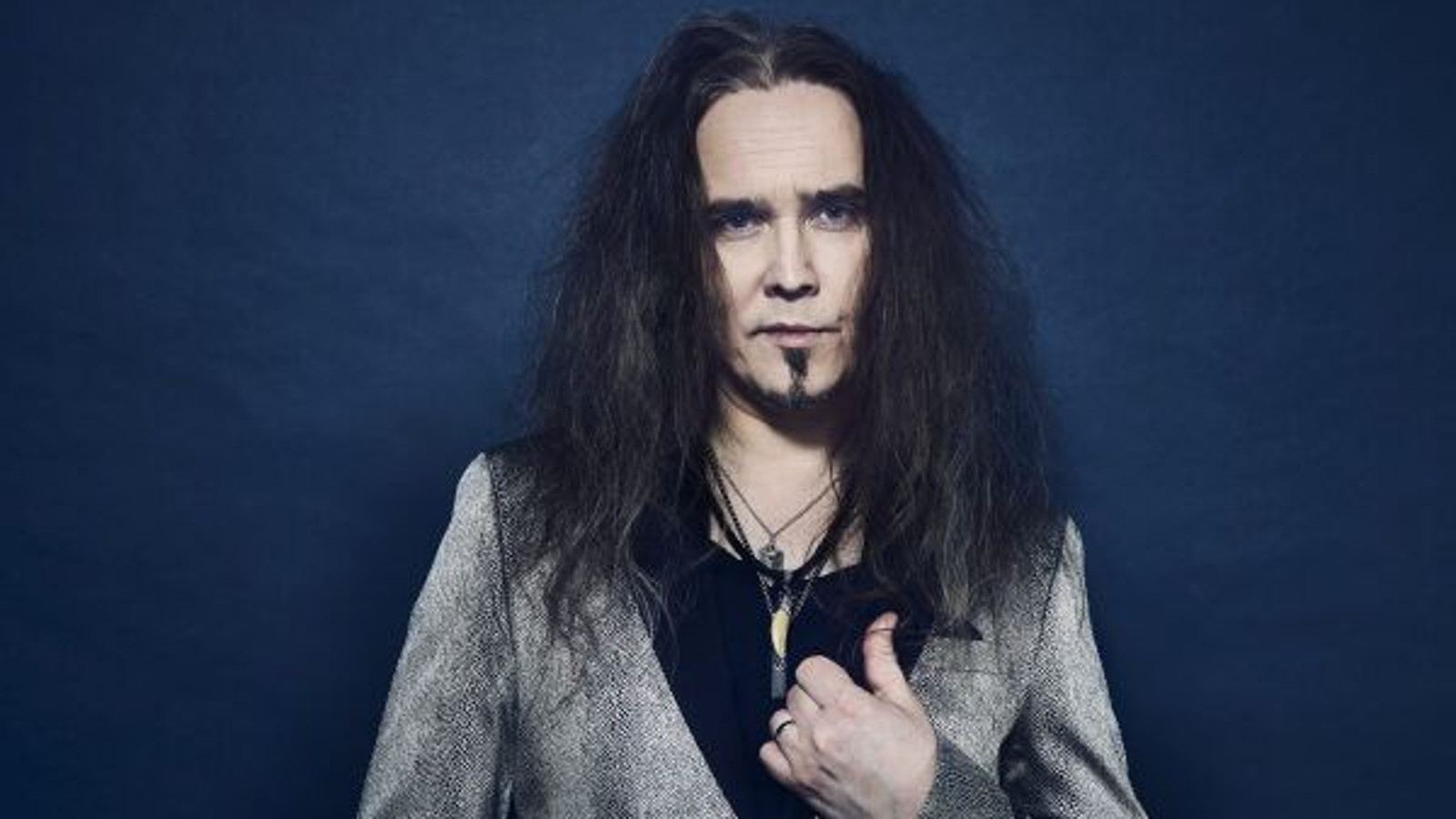 Kuvassa on Jarkko Ahola seisomassa ja hänellä on harmaa takki ja musta t-paita.  Tausta on tumman sininen.