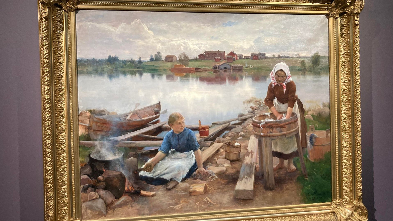 Kuvassa on kullan värisissä kehyksissä teos, jossa on järven rannalla nainen pesemässä pyykkiä korkean paljun ääressä.  Vieress vasemmalla on maassa istumassa tyttö, joka on keittopadan vieressä. Taustalla on tyyntä järveä, jonka takaa näkyy metsää ja taloja.