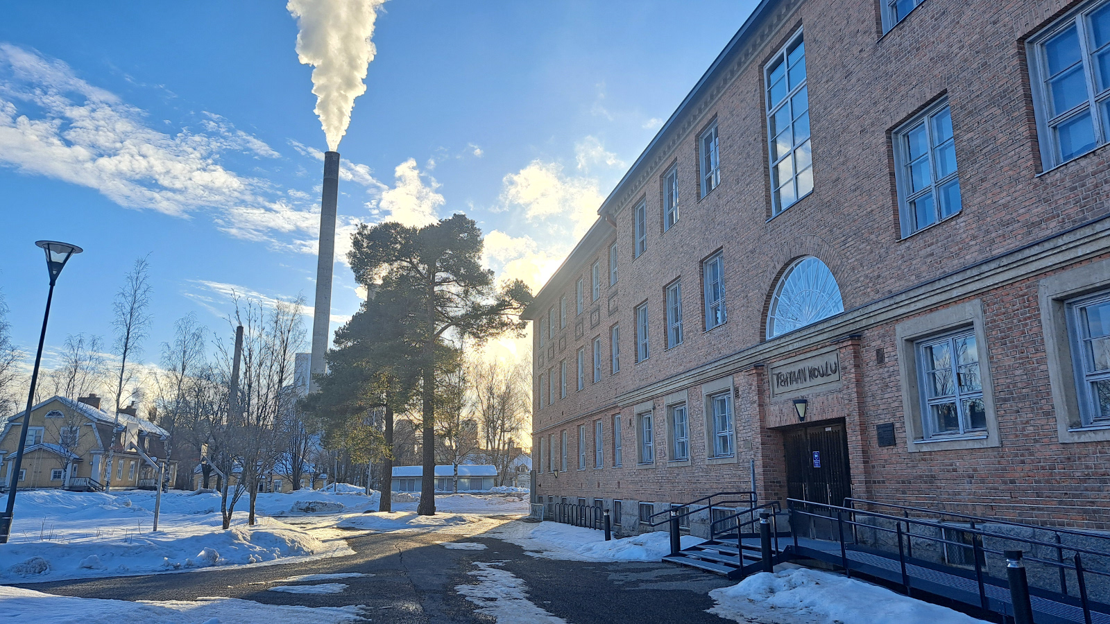 Kuvassa on Varkauden museokeskus Konstan vaaleata seinää oikealla vinottain ja siinä okolme kerrosta ikkunoita. taustalla näkyy männynn takana savuava, korkea tehtaan piippu seka talo. Etualalla on valkoista lunta ja kuvassa on pakkaskeli.