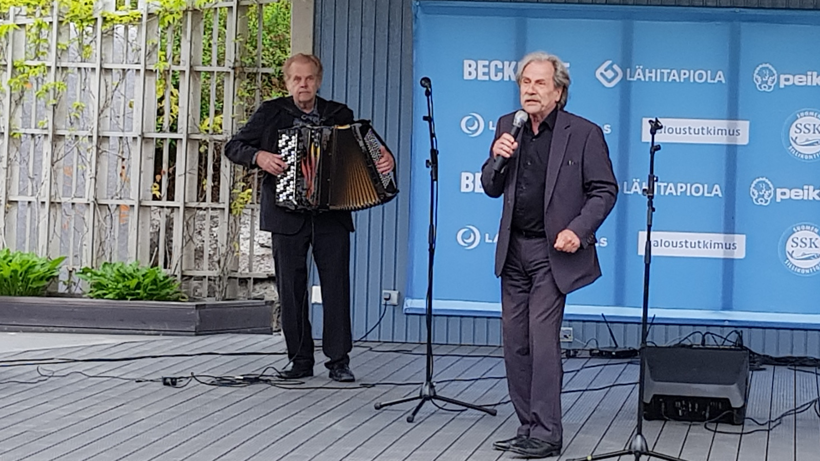 Kuvassa on Salmelan Puistonäyttämöllä vasemmalla hanuria soittamassa Heimo Mäntynen tumman harmaassa puvussa ja oikealla Erkki Liikanen myös tumman harmaassa puvussa.