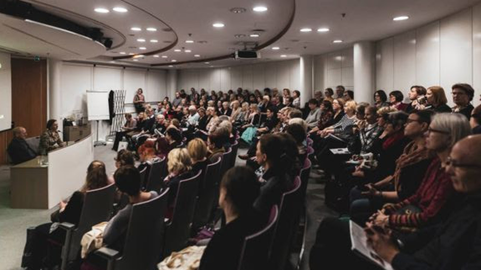 Kuvassa on auditorio, jossa on vasemmalla korkeammalla tasolla olevan pöydän ääressä nainen puheenjohtajana.  Hänen edessään on puolikaaressa yleisöä.