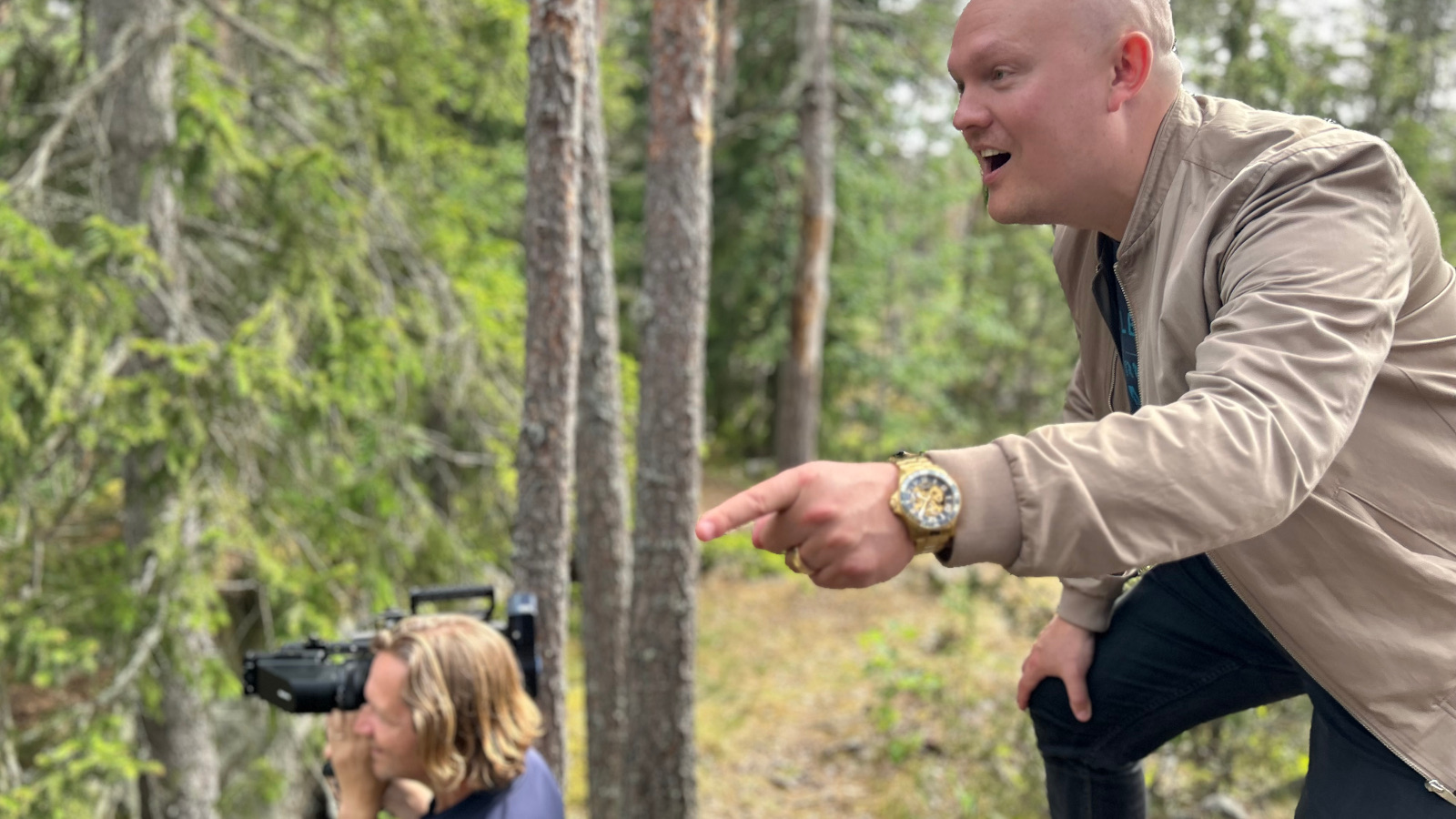 Kuvassa on sivuttain ylhäällä kallion päällä havumetsässä Samuel Harjanne ja osoittaa vasemmalla kädellään ja etusormellaan eteenpäin. Alempana on kuvaaja, josta näkyy pää ja kamera kuvaustilanteessa. 