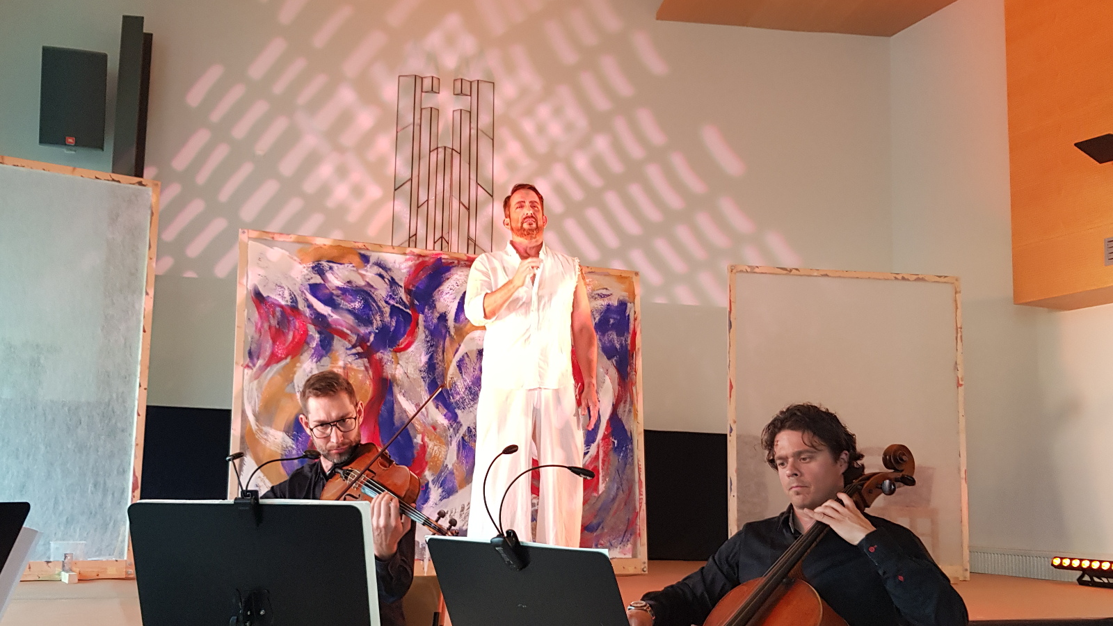 Kuvassa on keskellä valkoisessa asussa Kevin Greenlaw ja hänen takanaan on aaltoileva, valko-sini-punainen taulu. Edessä ovat viulisti Rèmi Moingeonja sellisti Samuli Peltonen mustissa vaatteissa edessään nuottitelineet.