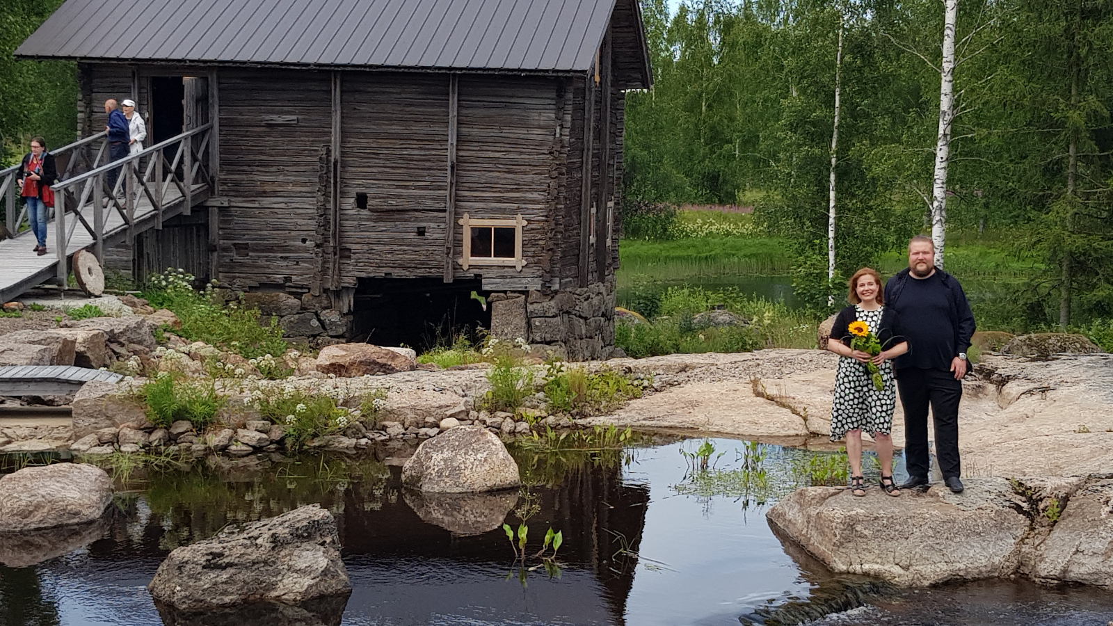 Kuvassa on vasemmassa kulmassa harmaa hirsimylly ja sen sillalla kulkee ihmisiä.  Edustalla on kalliota ja sen edessä virtaa vettä ja on kiviä  Oikealla alhaalla seisovat Erik Rousi ja Liisa Malmivaara, jolla on auringonkukka kädessään. 