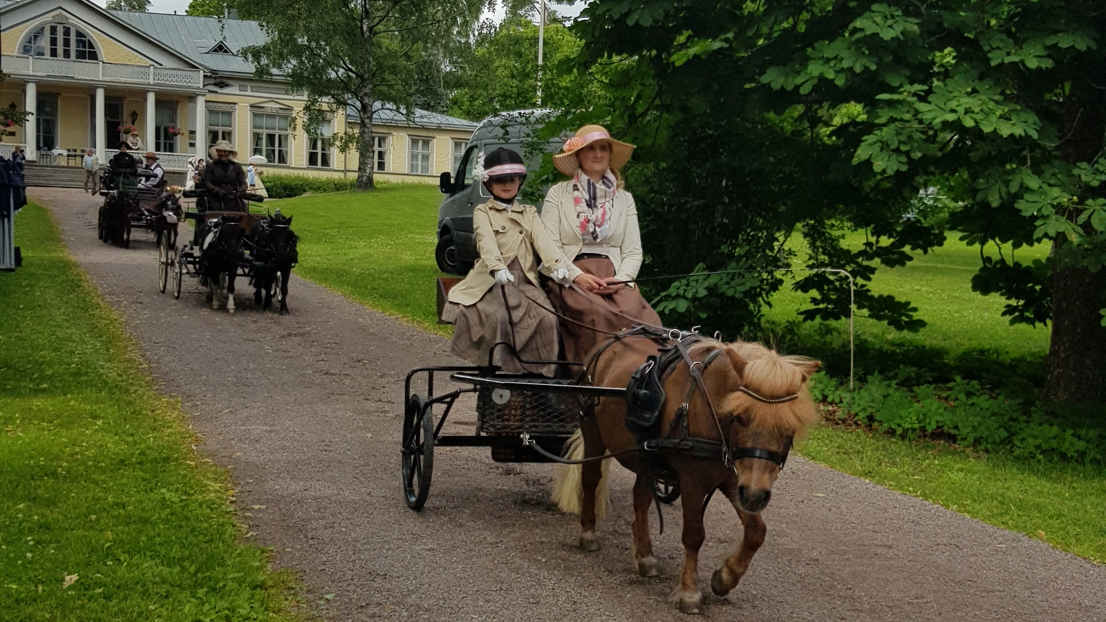 Kuvassa on tietä pitkin kolmet ponirattaat ja taustalla näkyy vaalea Harjun päärakennus. Oikealla on vanha lehmus.