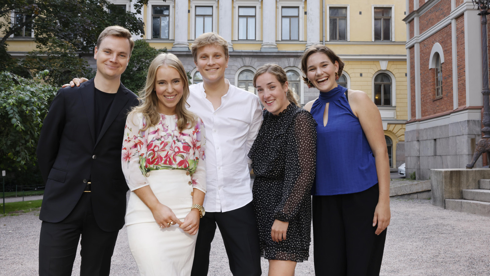 Kuvassa on ryhmä nuoria ja vasemmalla on mies mustissa vatteissa. Vieressä on nainen punakukallisessa paidassa valkoisessa puolihameessa. Kesekellä on mies valkosiessa paidassa ja vieressä nuori nainen tumman sinisessa mekossa ja toinen sinisessä puserossa. Taustalla on kelta-valkoinen rakenus klassisine pylväineen.