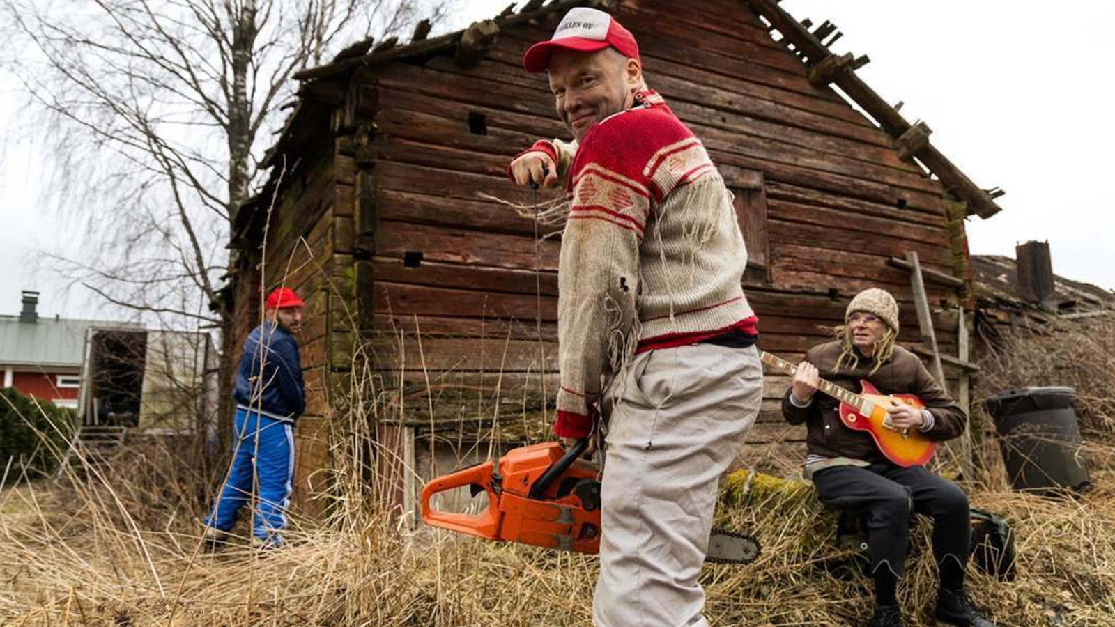 Kuvassa on nuori mies vetämässä moottorisahaa käyntiin ja hänellä on pohjalainen Jussi-paita päällään. Päässä on lippalakki. Taustalla on ruskea huonokuntoinen hirsiaitta ja sen vieressä selin pissalla tumman sinisessä pusakassa ja sinisissä verkkareissa toinen mies. Kannon päällä oikealla on nuori mies vaaleassa pipossa soittamassa kitaraa.