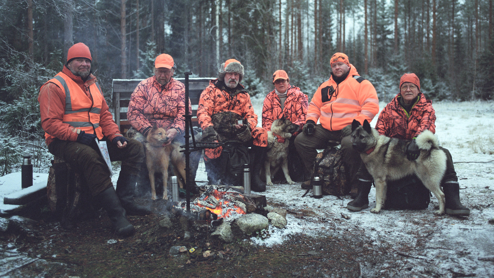 Kuvassa on miehiä nuotion äärellä punaisissa metsästyspusakoissa ja oikealla edessä on harmaa koira.  Maassa on jonkin verran lunta.