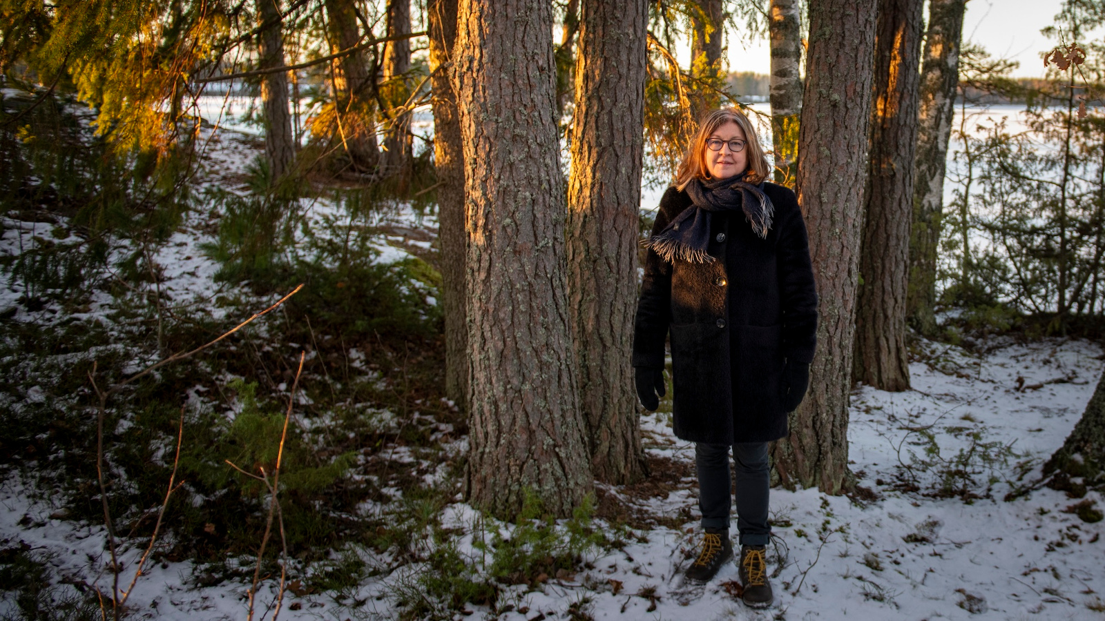 Kuvassa on Eija-Liisa Ahtila seisomassa kuusi- ja mäntymetsikössä isojen männyn runkojen vieressä.  Maassa on hiukan lunta. Hänellä on musta takki päällään.