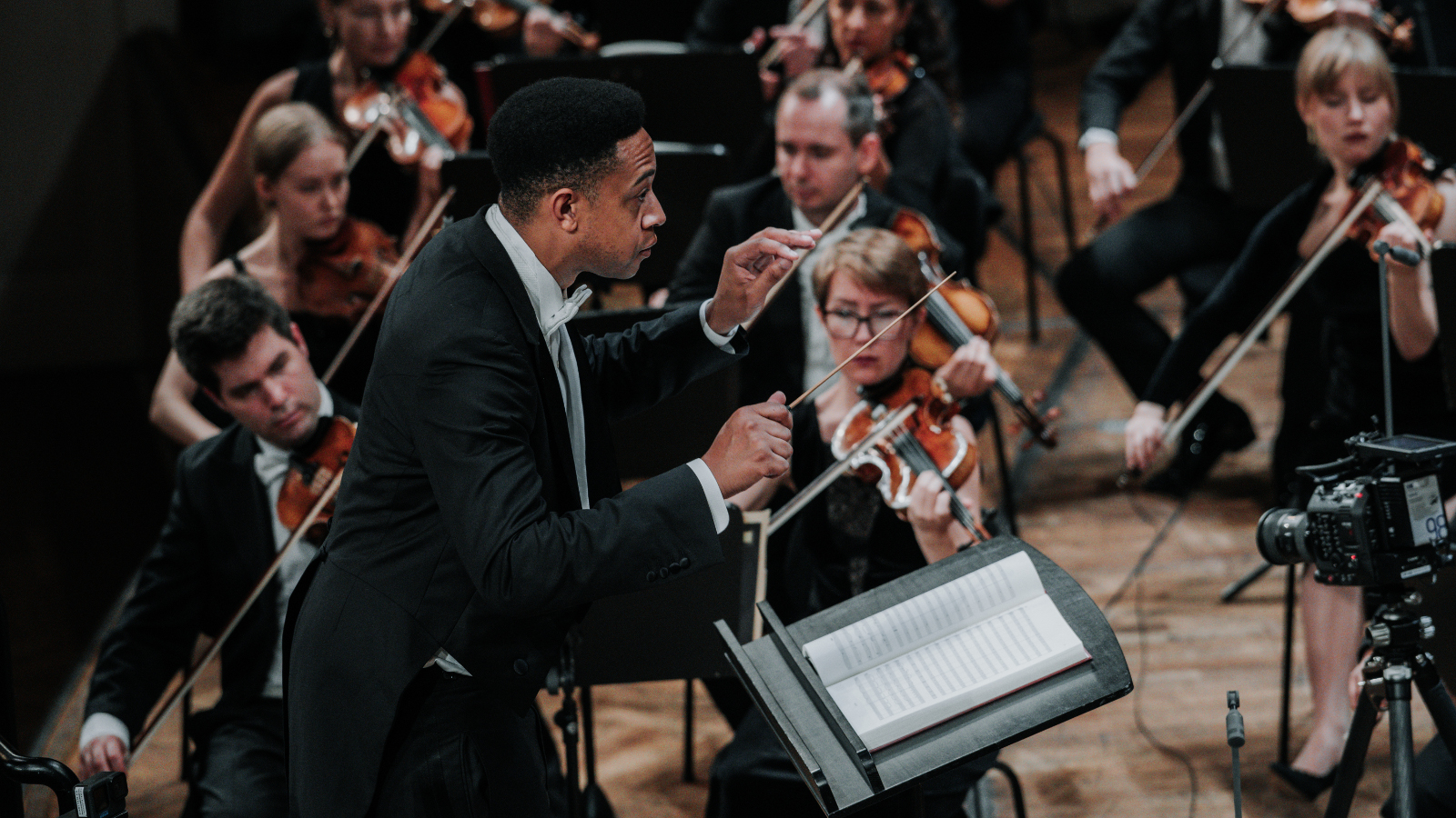 Kuvassa on Nova Orchester Wienin johtaja Wsivuttain seisomassa ja johtamassa orkesteria. Orkesterilaisia näkyy hänen takanaan soittamassa.