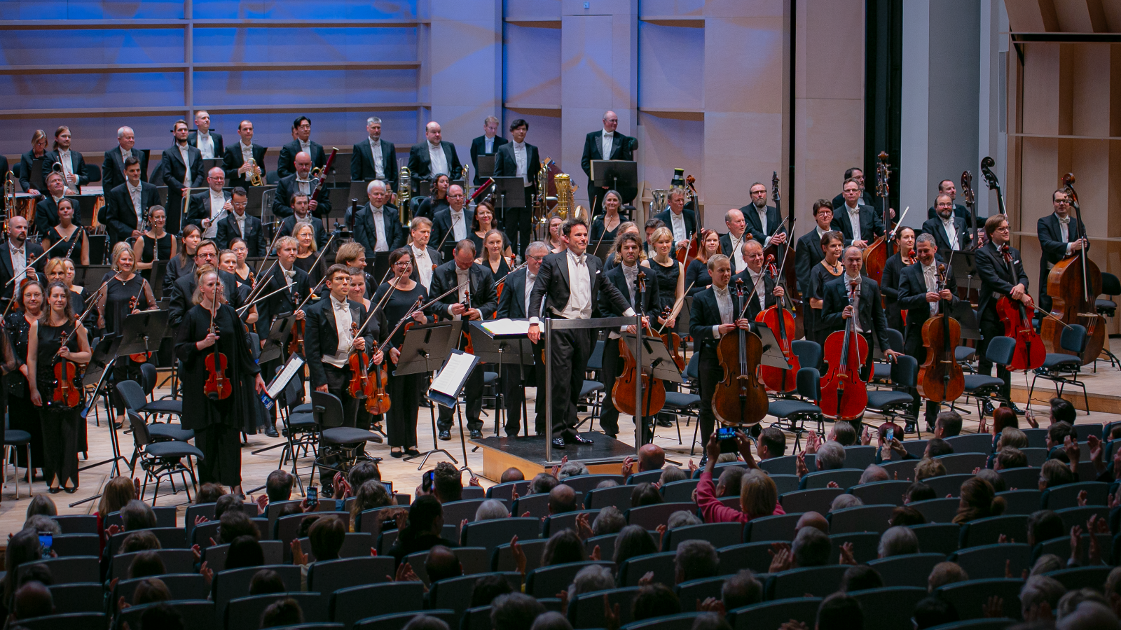 Kuvassa on lavalla Tampere Filharmonian soittajat seisomassa soittimineen. Edessä on yleisöä istumassa.