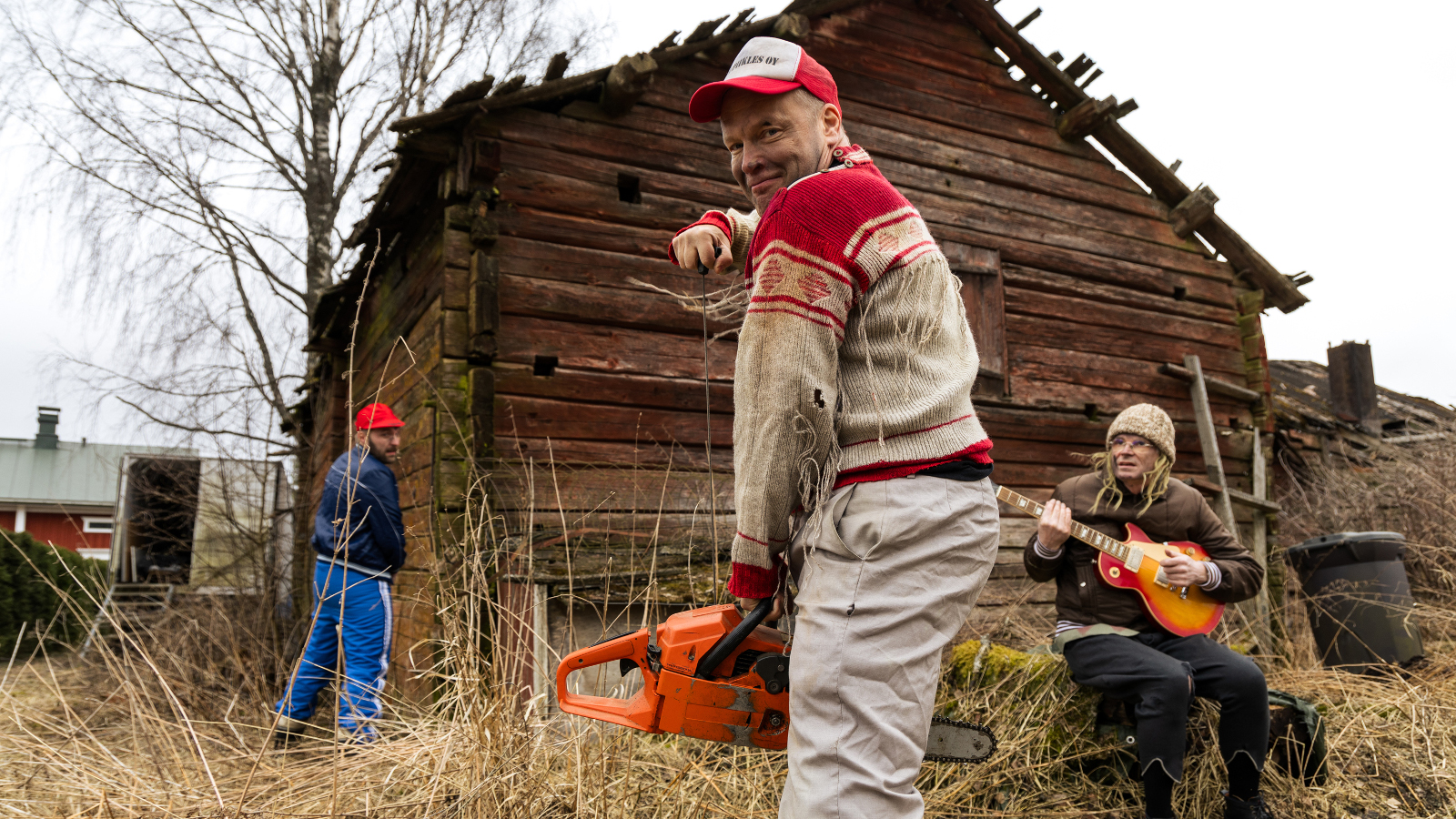 Kuvassa on keskellä ruskea hirsinen riihi ja pitkäruohoista maalaispihaa. Etualalla seisoo mies, jolla on Jussi-paita päällään ja vaaleat housut.  Hänellä on oranssi moottorisaha kädessään.  Hänen takanaan on mies soittamassa mandoliinia kannon päällä. Riihen vieres on selin mies sinisessä verkkarissa.