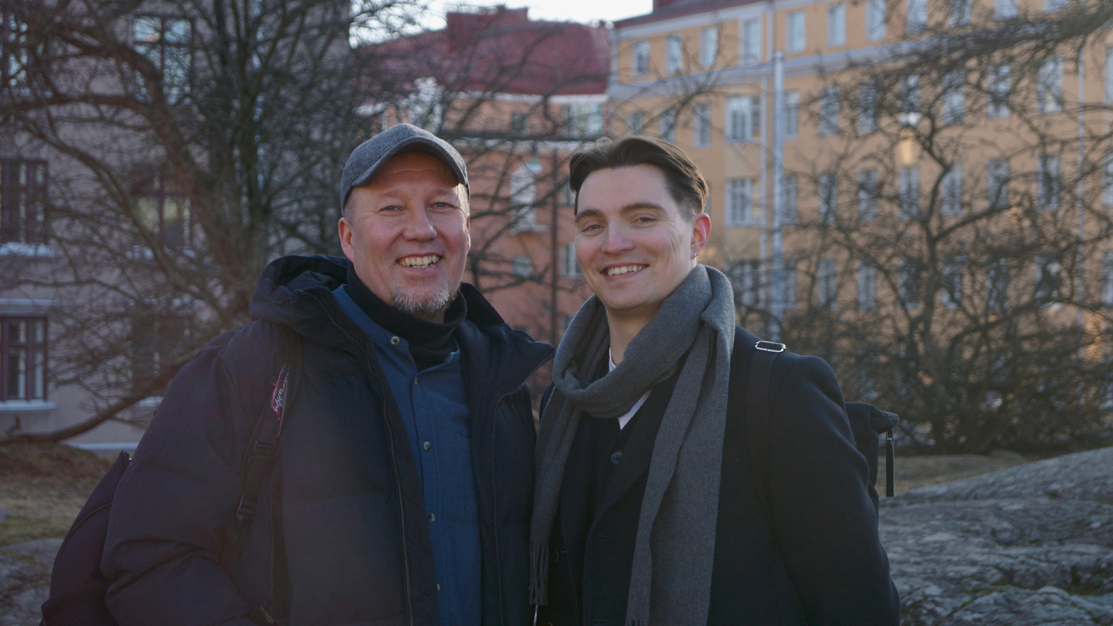 Kuvassa ovat Mikko Korsulainen vasemmalla tummassa takissa ja lippalakissa.  Oikealla on Rasmus Arikka mustassa takissa ja paljain päin.  Molemmat ovat puolivartalokuvassa ulkona ja taustalla näkyy paljaita lehtipuun runkoja ja oikealla on vaalea kerrustalo. 