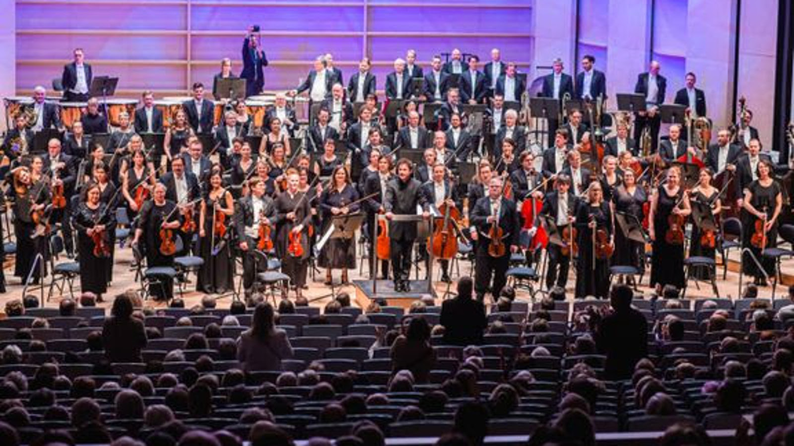 Kuvassa on Tampereen Filharmonia seisomassa lavalla soittimet kädessään ja mustissa puvuissa. Etualalla on penkkiriveissä yleisöä ja osa seisomassa.